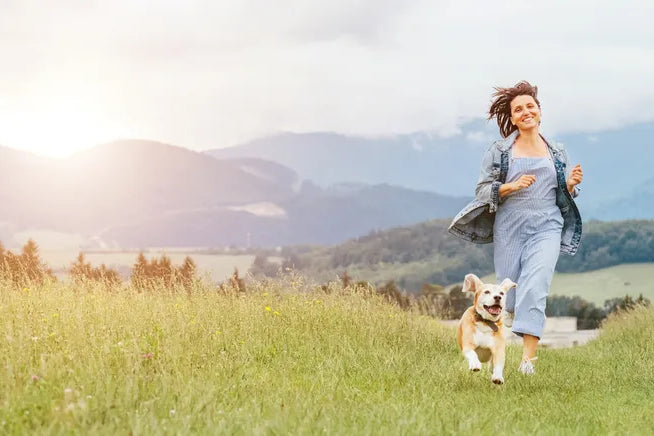 Frau rennt lachend mit Hund über Wiese, symbolisiert Loslassen und Neuanfang in der Natur.