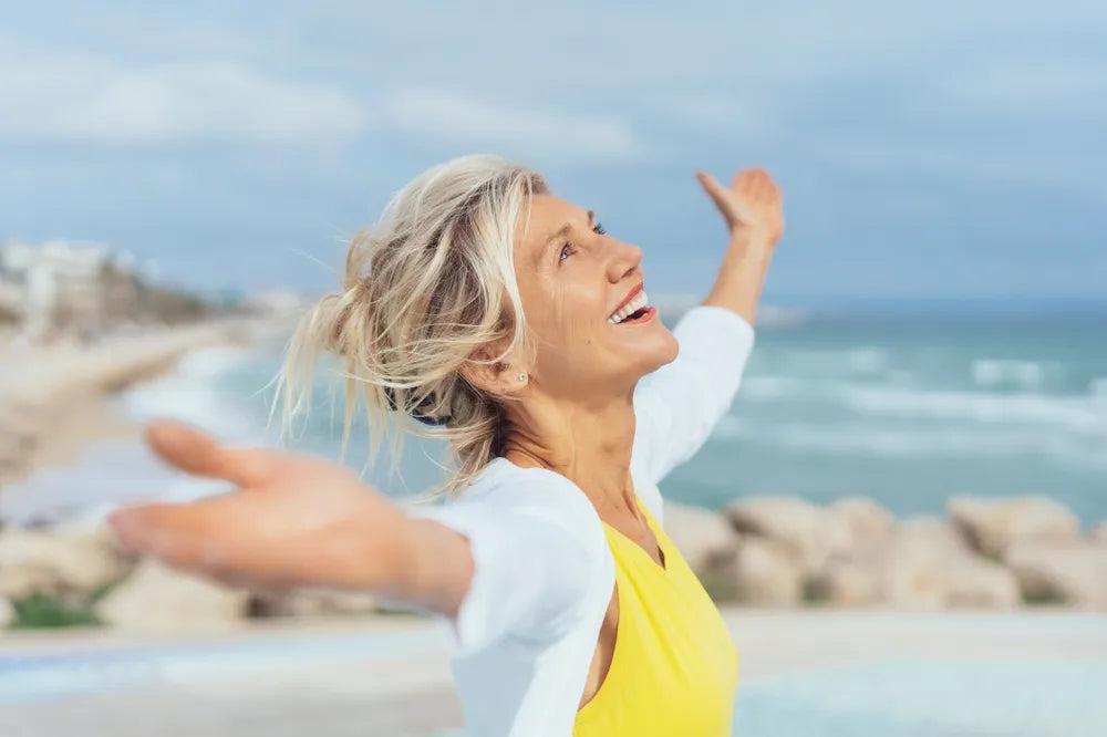 Frau mit ausgebreiteten Armen am Meer, lacht, symbolisiert positive Affirmationen und Freiheit.