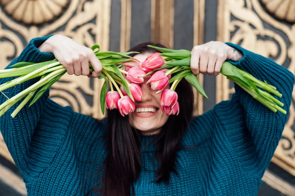 Frau hält rosa Tulpen vor das Gesicht und lacht, symbolisiert Selbstliebe und Freude.