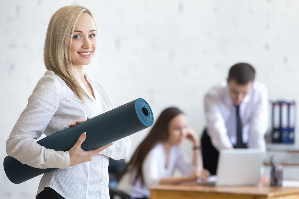 Junge Frau hält Yogamatte im Büro, lächelt, symbolisiert Stressbewältigung und Entspannung.