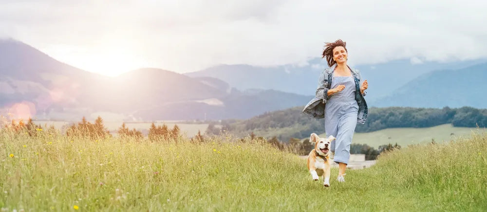 Frau rennt lachend mit Hund über Wiese, symbolisiert Loslassen und Neuanfang in der Natur.